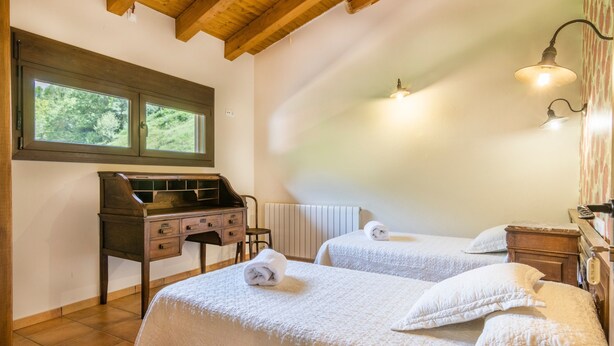 Une chambre avec deux lits, un bureau en bois, un radiateur et une fenêtre donnant sur des arbres.