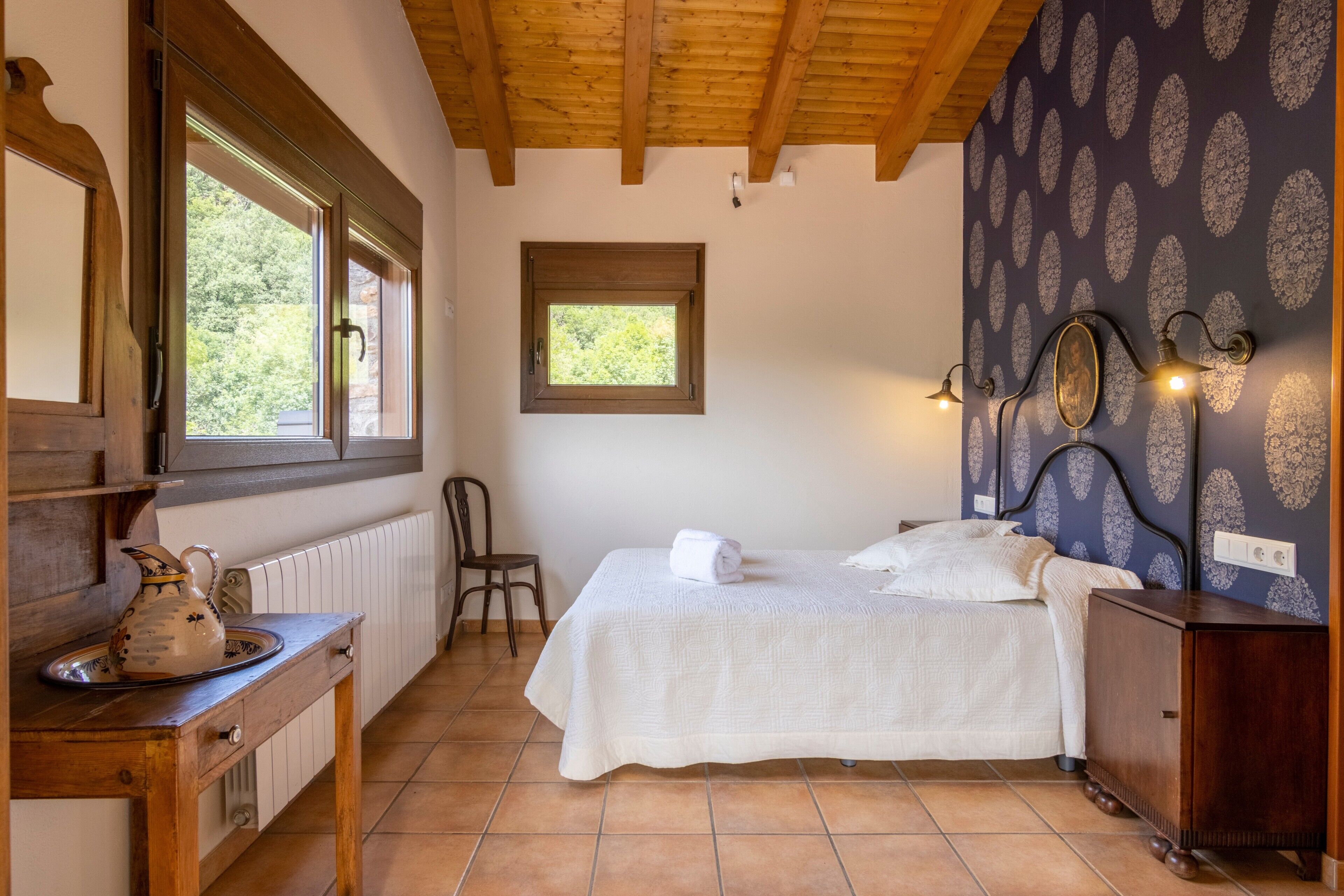 Un dormitorio con cama, cómoda de madera, una silla, ventana y pared con estampado.