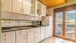 Une cuisine avec des armoires blanches, un comptoir noir, un évier en acier inoxydable, un four à micro-ondes et un réfrigérateur. Une porte-fenêtre coulissante donne accès à un espace extérieur avec vue sur un village.