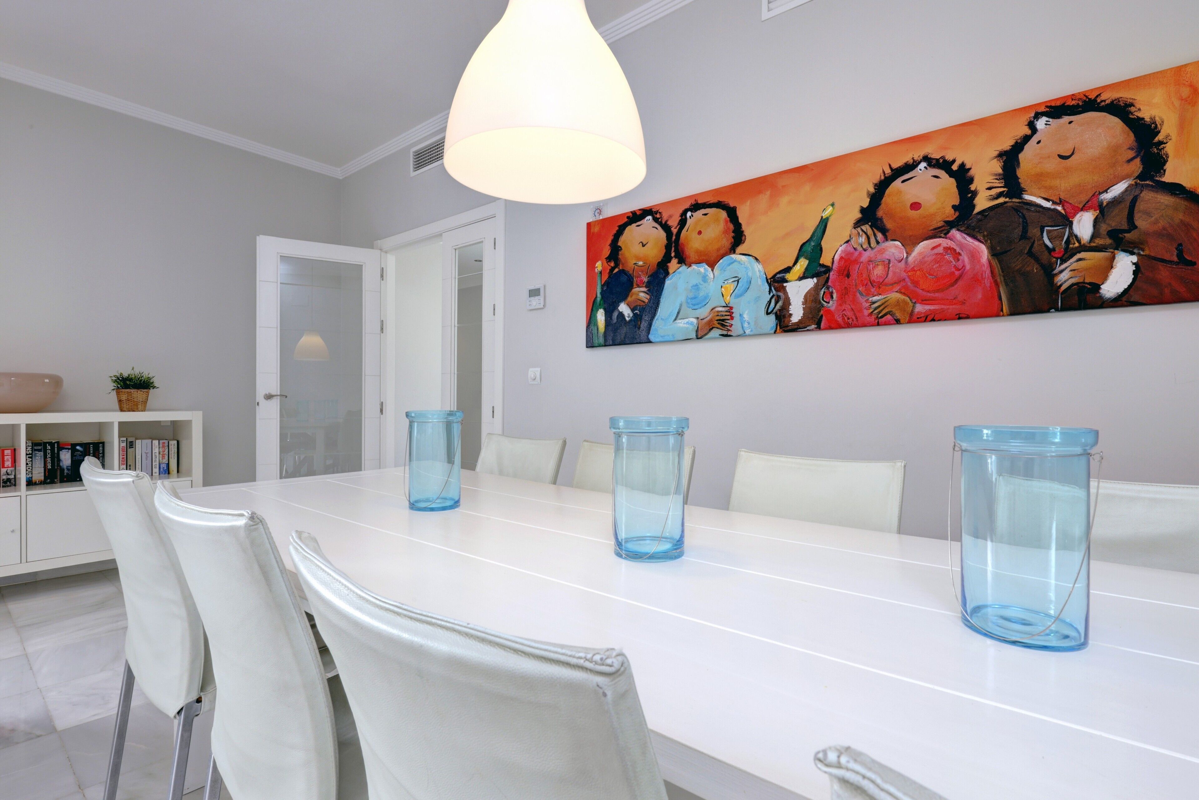 A dining area with a white table, blue glass candle holders, white chairs, a painting on the wall, and a white cabinet with books.