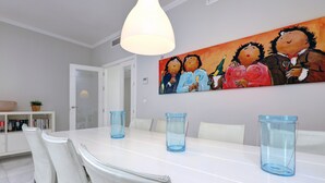 A dining area with a white table, blue glass candle holders, white chairs, a painting on the wall, and a white cabinet with books. - Apartment "Mirador de la Cala" with Terraces & Wi-Fi (Estepona)