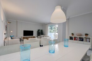 A modern living room with a white sofa set, a large flat-screen TV, a dining table with blue glass vases, and a bookshelf with books and decorative items.