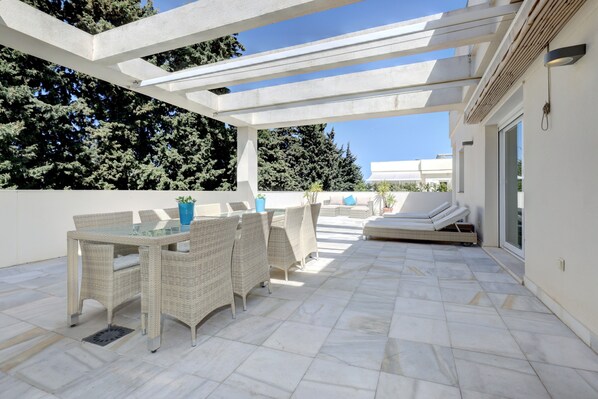 An outdoor patio with wicker furniture, a glass table, and a white ceiling structure.
