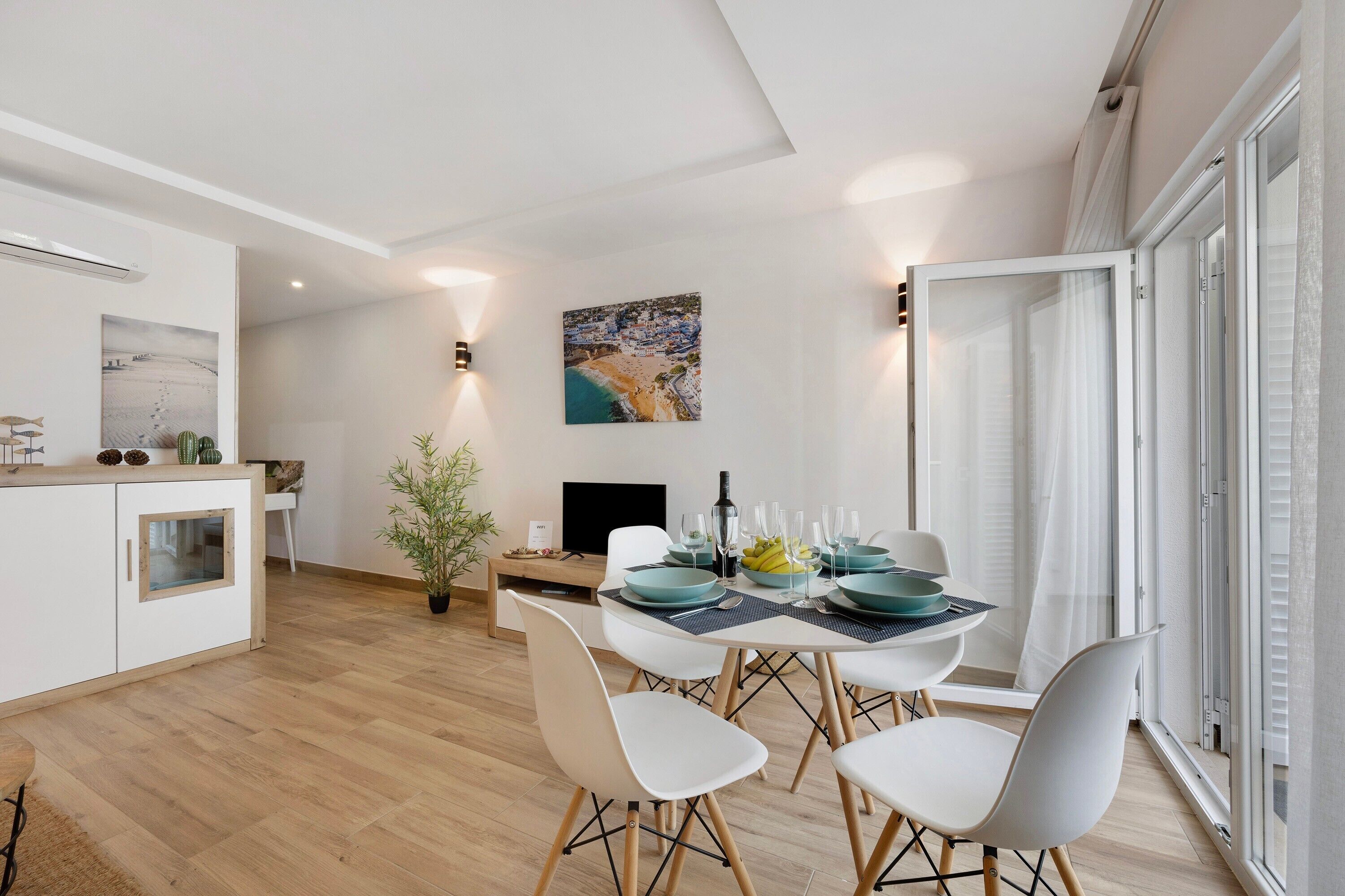 Una cocina moderna con armarios blancos, un comedor con mesa para cuatro personas y un gran ventanal con vista a la playa.
