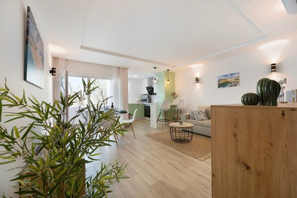 A modern living room with a sofa, a dining area with chairs, a kitchen in the background, and a large plant in the foreground.