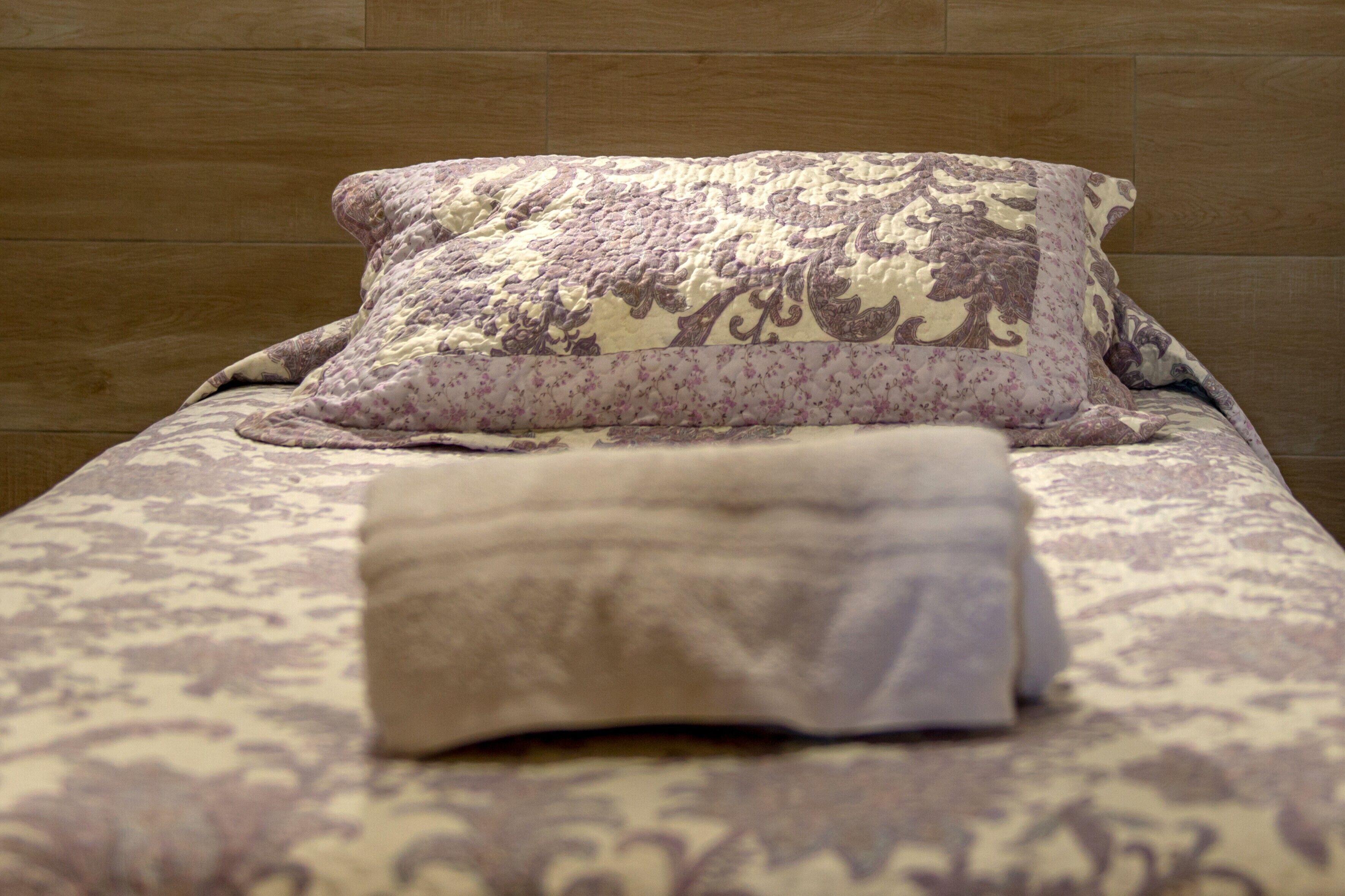 Una cama con una colcha estampada y almohadas, una manta doblada y paneles de madera en la pared.