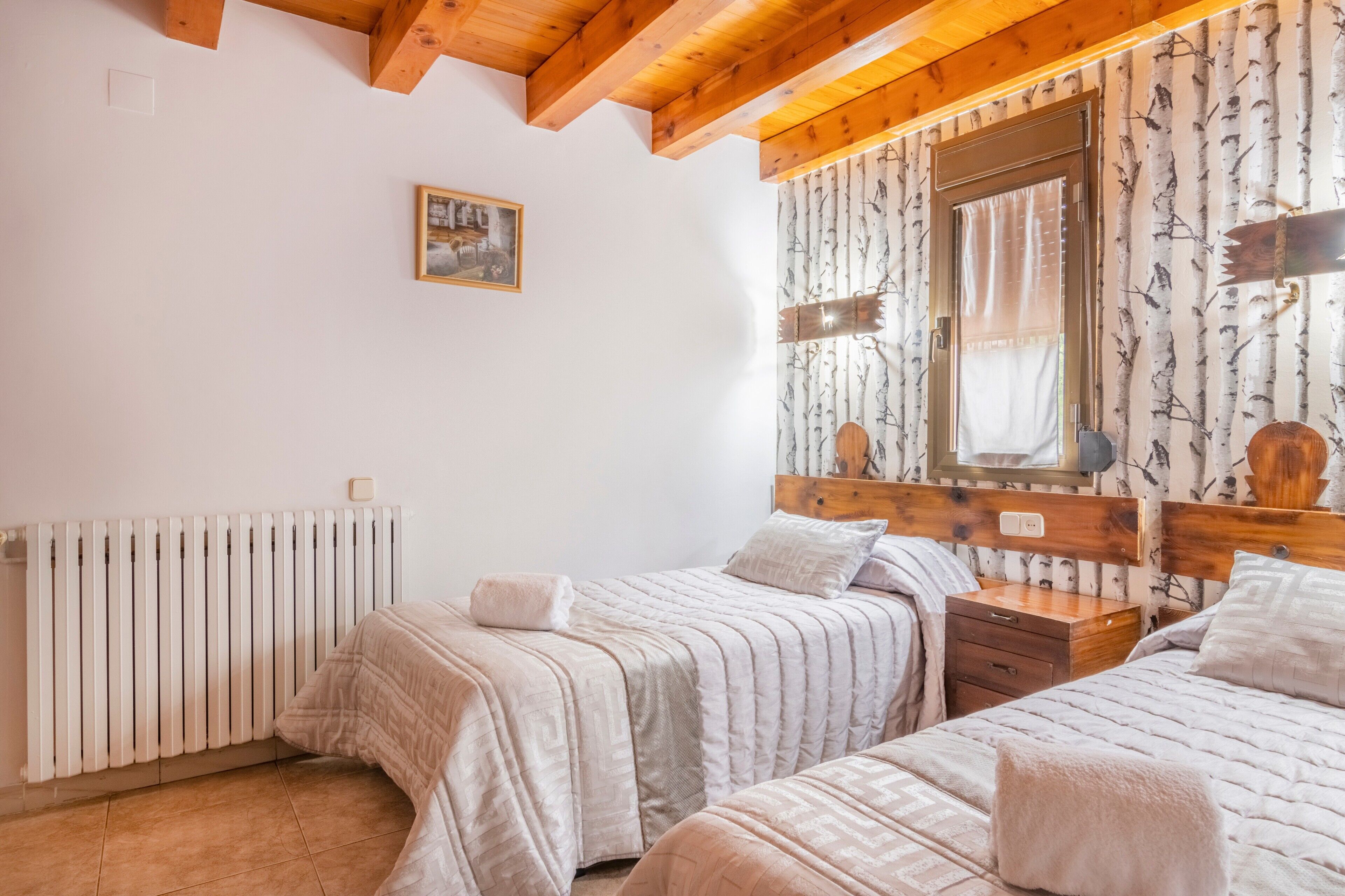 Un dormitorio con dos camas, techo de madera, radiador, ventana con cortina y un espejo fijado a la pared.