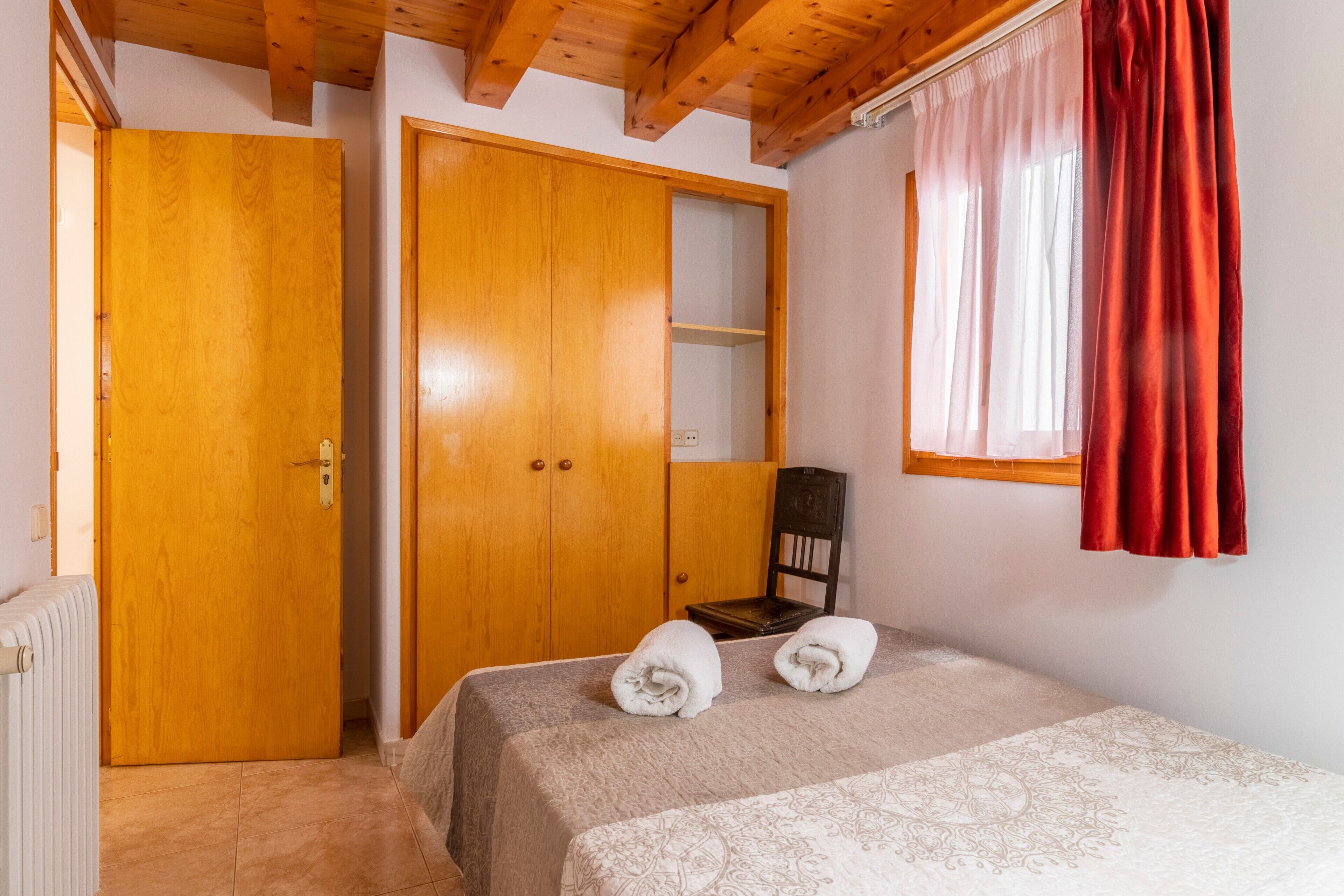 Een slaapkamer met een bed, twee houten kasten, een radiator en een raam met rode gordijnen.
