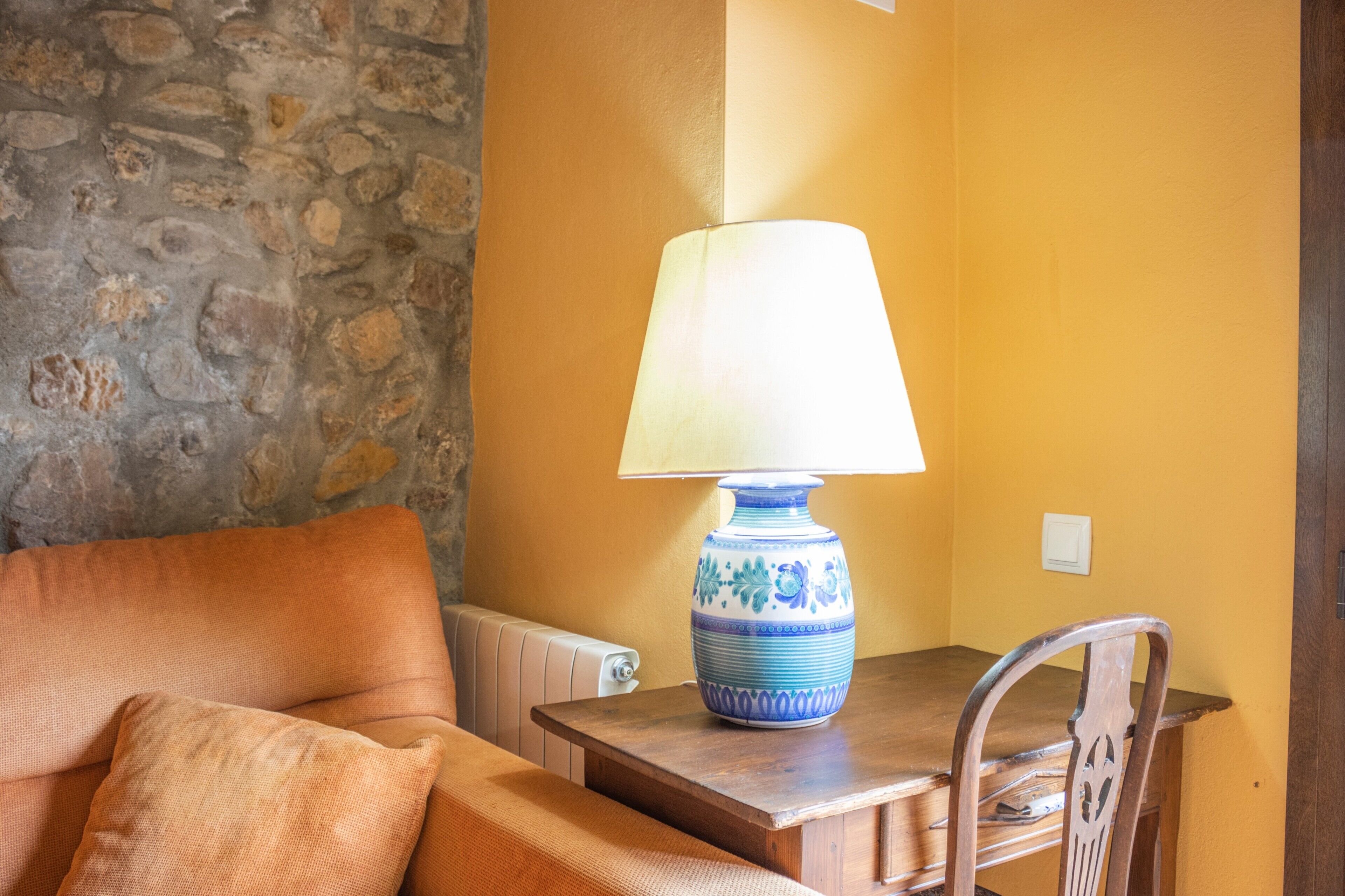 Une pièce avec un mur en pierre, une table en bois avec une lampe, une chaise et un radiateur.