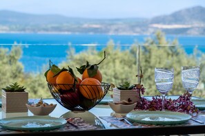 A table with a fruit bowl, water glasses, and plates, overlooking a scenic view of a body of water and greenery.