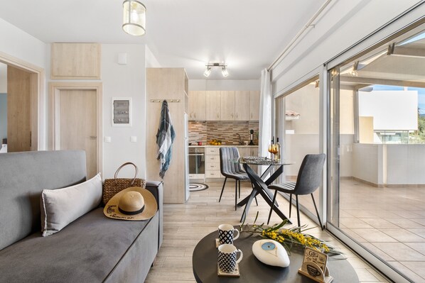 A modern living room with a sofa, a dining table with chairs, a kitchen area, and a balcony with a view.