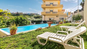 A poolside area with lounge chairs, a building with balconies, and a clear blue sky. - Apartment 'Rafaela's House' with Shared Pool, Wi-Fi and Air Conditioning (Koskinou)