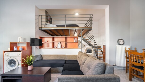 A modern living room with a sectional sofa, a flat-screen TV, a washing machine, and a staircase leading to an upper level. - Apartment 'Rafaela's House' with Shared Pool, Wi-Fi and Air Conditioning (Koskinou)