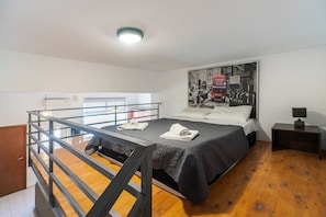 A bedroom with a bunk bed, a bed with a dark blanket, a poster on the wall, a nightstand, and a wooden floor.