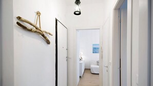 Een hal met witte wanden, een houten vloer en een deur die leidt naar een slaapkamer met een bed en nachtkastje.