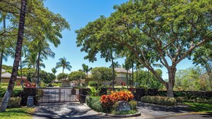 A gated driveway leading to a residential area with lush greenery and palm trees.