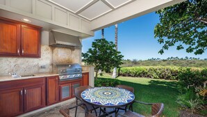 An outdoor kitchen with a round table and wicker chairs, surrounded by a lush garden and a clear blue sky.