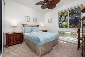 Un dormitorio con una cama, dos mesitas de noche, un aparador, un ventilador de techo y una ventana con persianas.
