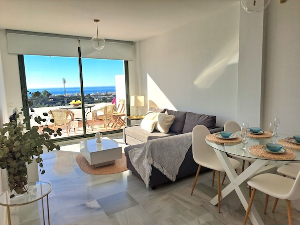 A modern living room with a sofa, dining table, and chairs, a balcony with a view of the sea, and a large window.