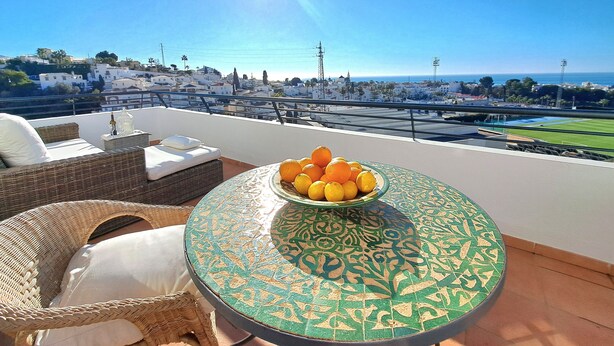 A balcony with wicker furniture, a table with a decorative tile, and a bowl of oranges. - Holiday Apartment "Paraíso Azul" with Sea View, Shared Pool (Summer) & A/C (Nerja)