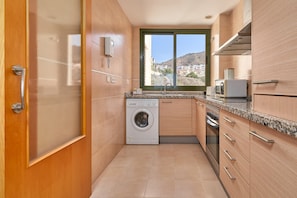 A compact kitchen with a built-in washer, a window with a view of a hillside, and wooden cabinetry.