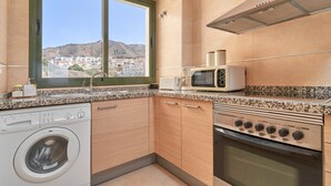 A modern kitchen with a washing machine, microwave, oven, and a view of a hillside town. - Holiday Apartment "Paraíso Azul" with Sea View, Shared Pool (Summer) & A/C (Nerja)