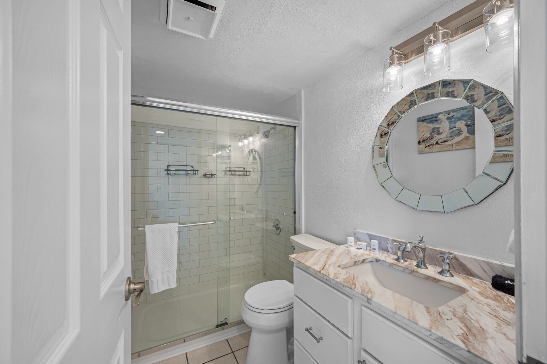 Une salle de bain avec une douche à parois de verre, des toilettes blanches, un lavabo en marbre et un miroir rond orné d’une peinture représentant une mouette.
