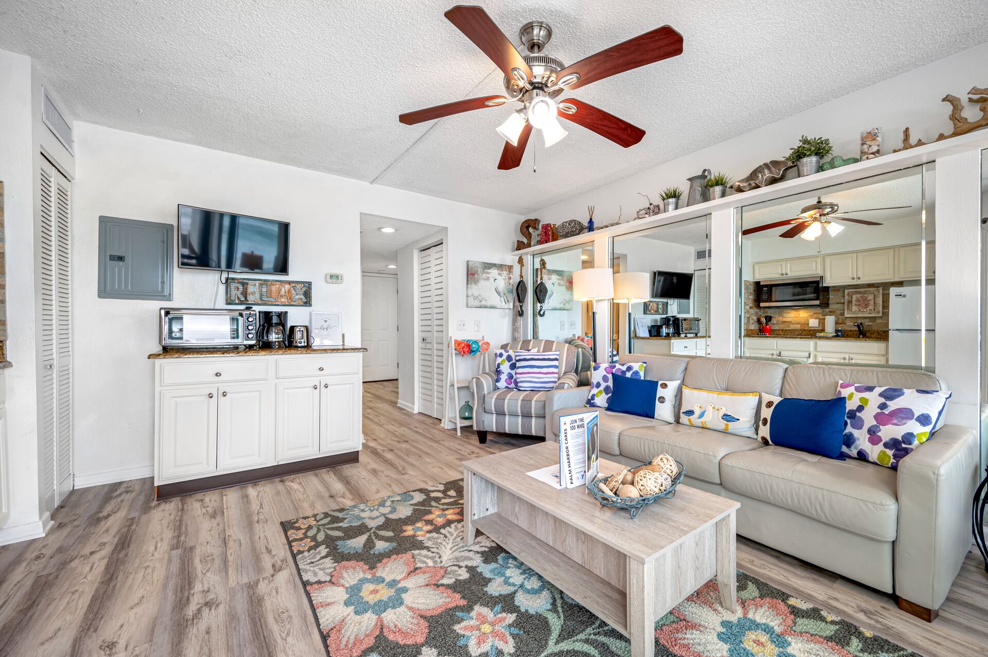 A modern living room with a sofa, coffee table, and a television. There is a kitchen area with cabinets and a microwave.