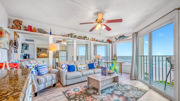 A spacious living room with a large window overlooking the beach, a ceiling fan, and a variety of decorative items on shelves.