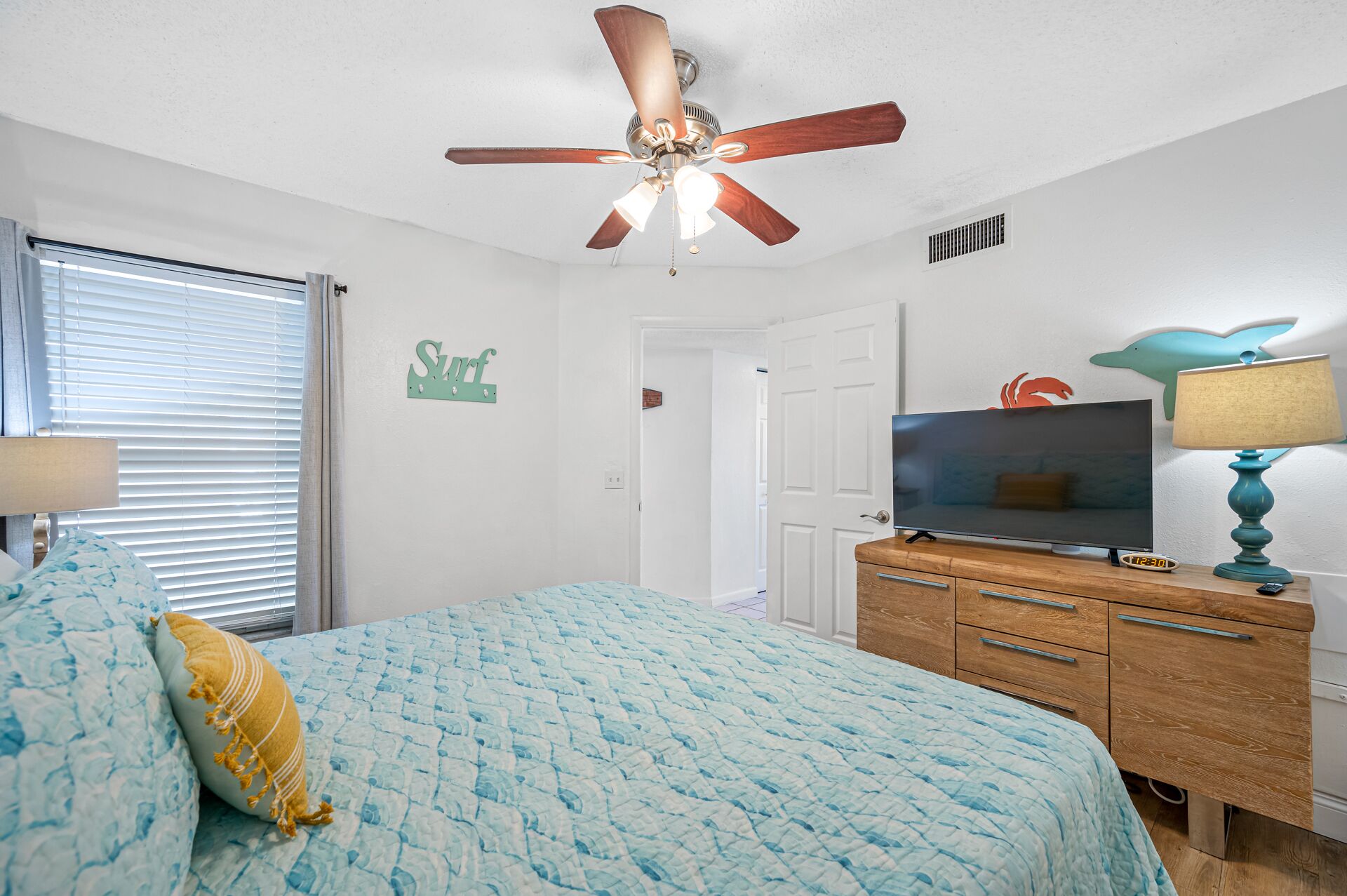 A bedroom with a bed, a ceiling fan, a TV, a dresser, and a wall with a 'Surf' sign.