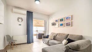 A modern living room with a grey sofa, a chair, and a table. There is a window with curtains, a wall-mounted air conditioner, and framed pictures. - City beach apartment (Chania)