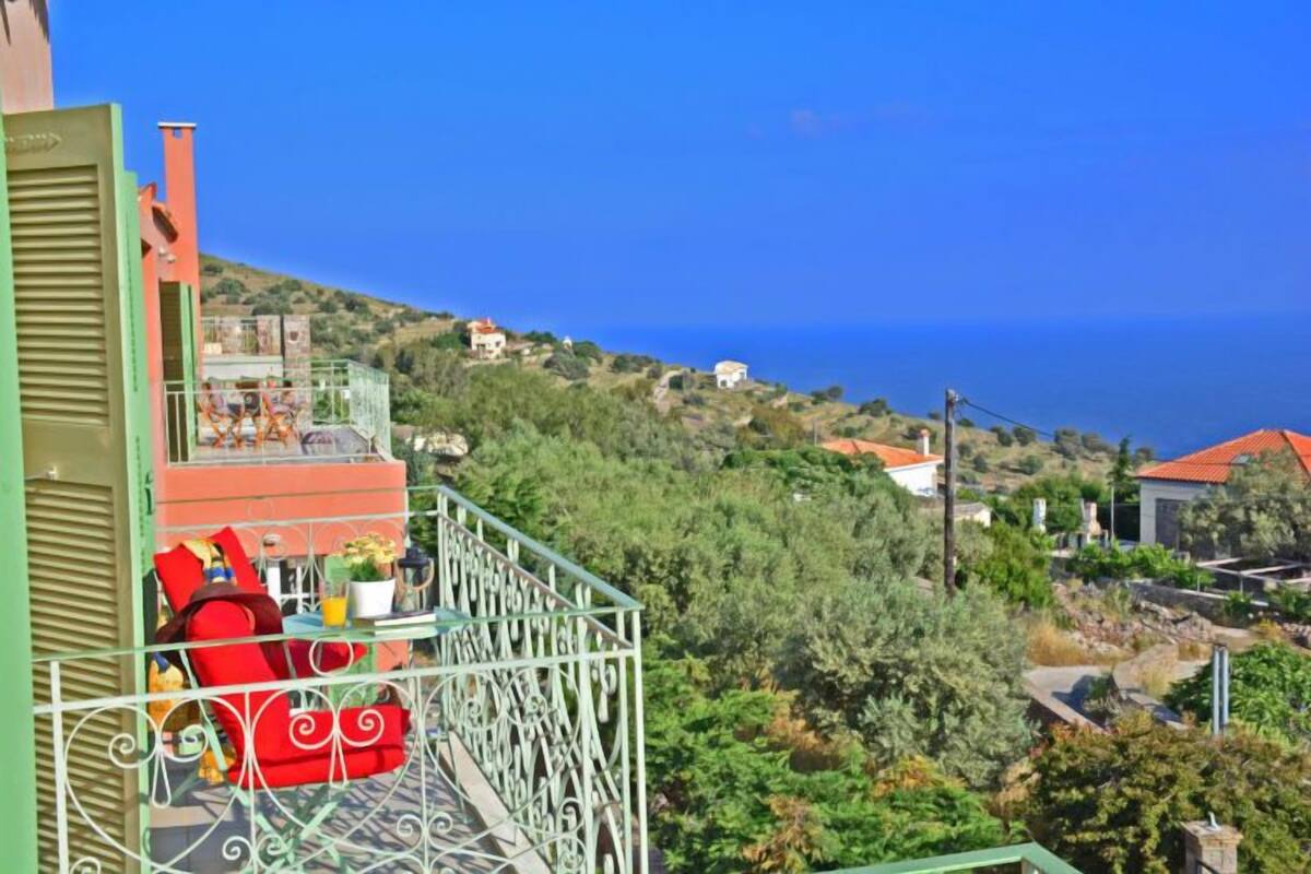 Un balcón con una silla y una mesa roja, con vistas a una ladera con edificios y un cielo azul despejado.