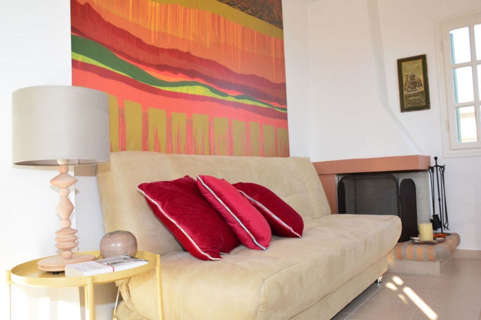 Una sala de estar con un sofá color beige, almohadas rojas, una mesa redonda con una lámpara y un cuadro abstracto de colores en la pared.