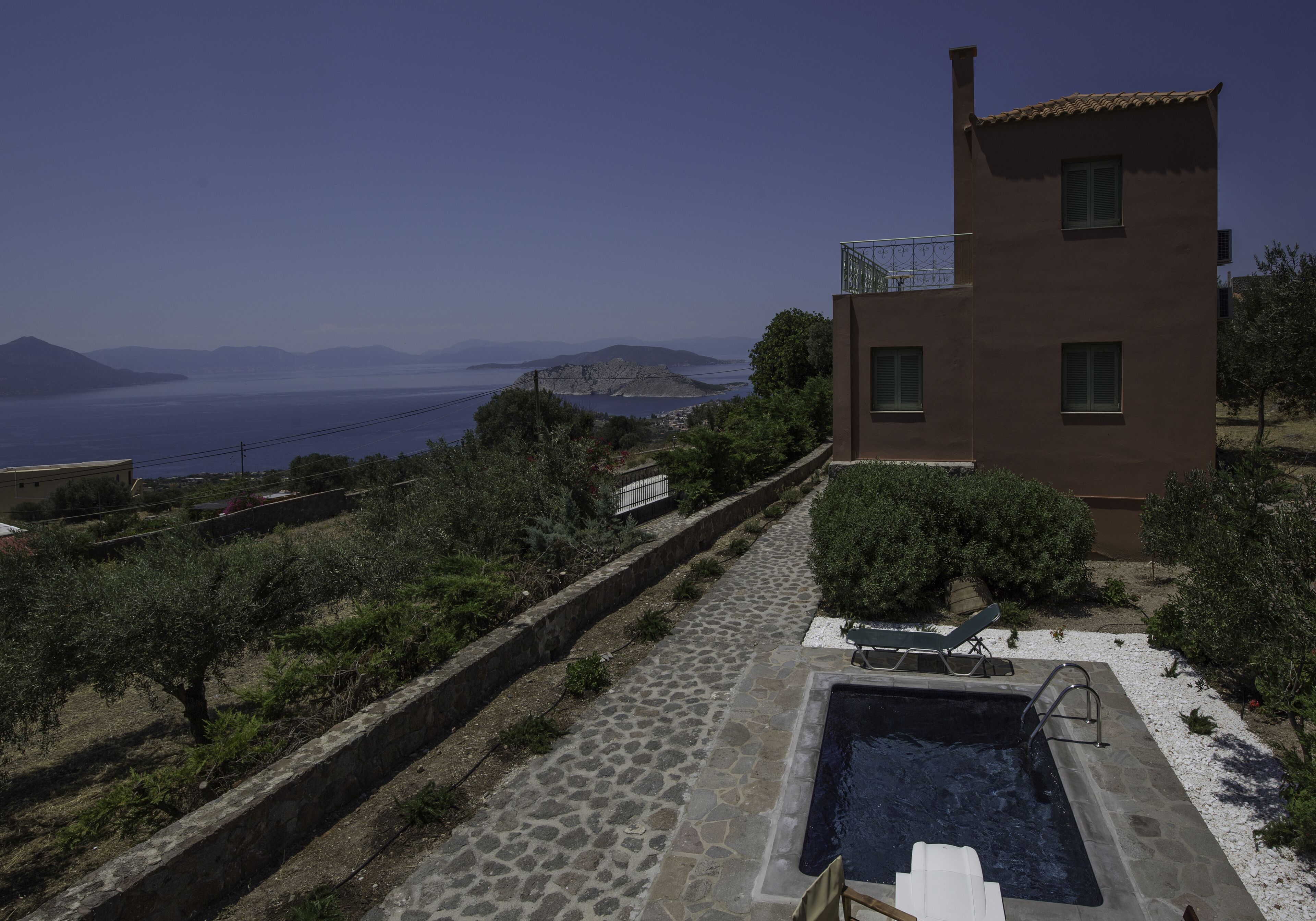 A modern villa with a stone-paved pool area, a small outdoor seating area, and a view of the sea and distant mountains.