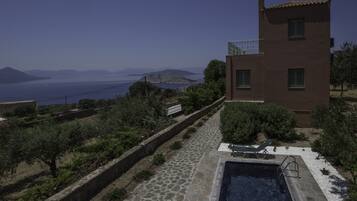 A modern villa with a stone-paved pool area, a small outdoor seating area, and a view of the sea and distant mountains.