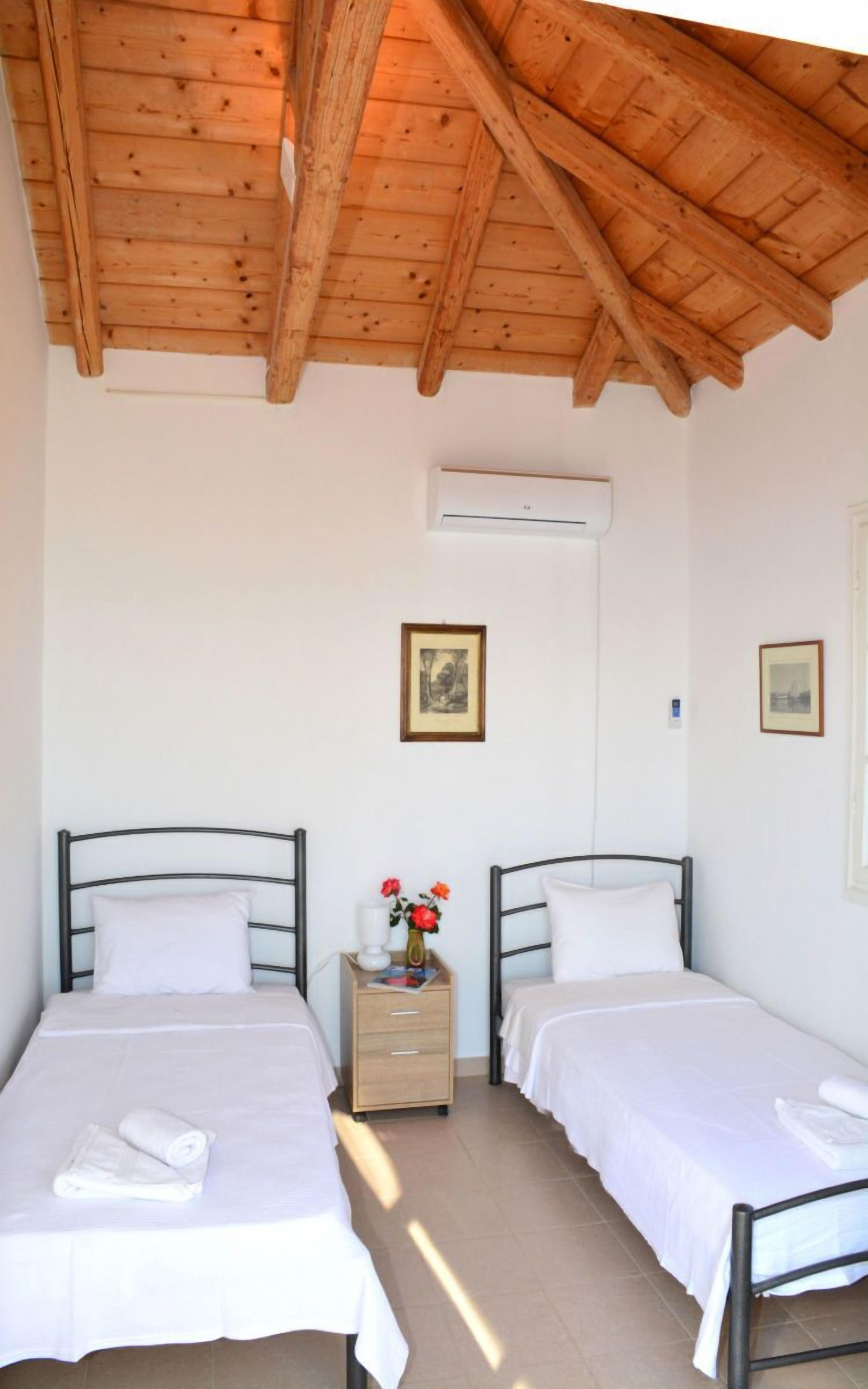 A room with two beds, wooden ceiling beams, an air conditioning unit, and a small bedside table with a vase of flowers.