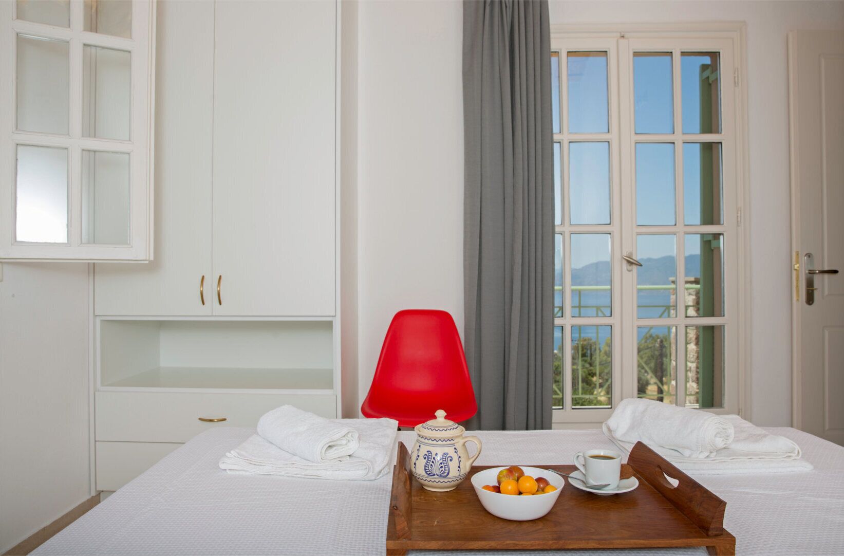 A hotel room with a bed, a table with a tray of fruit and a cup, a red chair, a window with a view of the sea, and a white wardrobe.