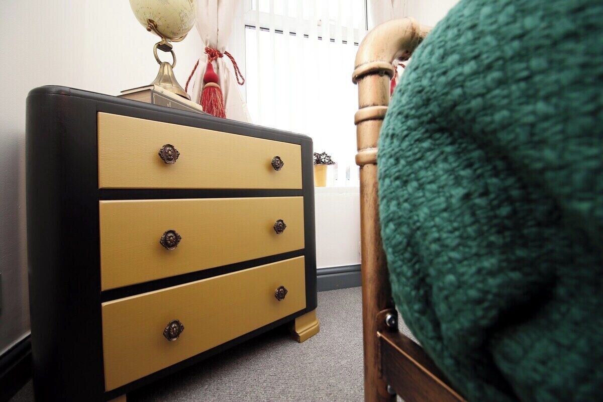 Een kamer met een houten stoel, een gele dressoir met drie lades, en een wereldbol erbovenop.