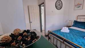 A bedroom with a bed, a bedside table with a bowl of dried flowers, a clock on the wall, and a door leading to another room. - Junctions Way by Tŷ SA -3 bed in Newport (Newport)
