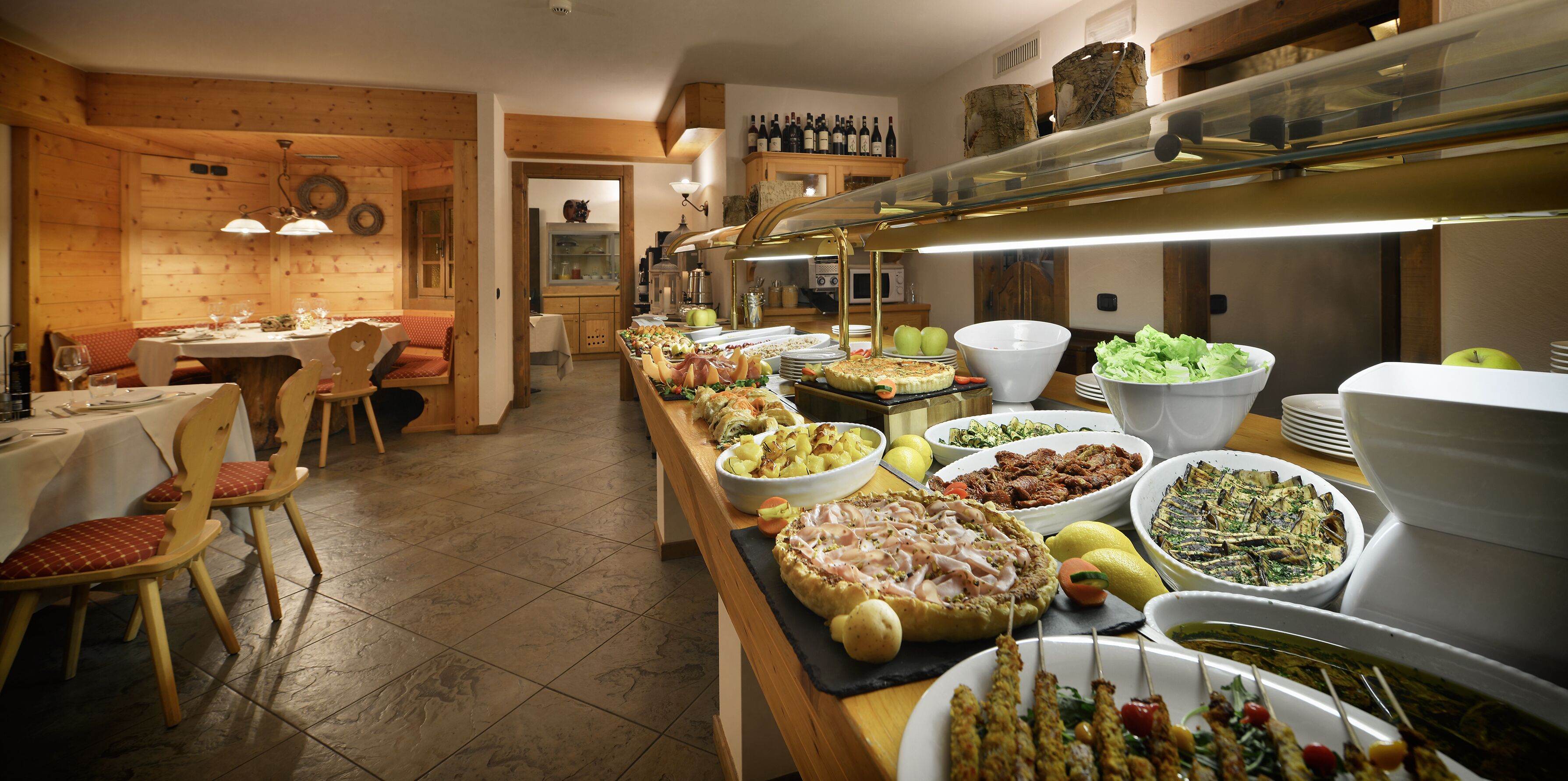 Een buffet met diverse gerechten, een eetgedeelte met tafels en stoelen, en een keukenruimte met wijnflessen en een koelkast.
