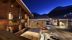 A wooden building with a balcony and a view of a snow-covered street and mountains at night. - Hotel Primula (Livigno)