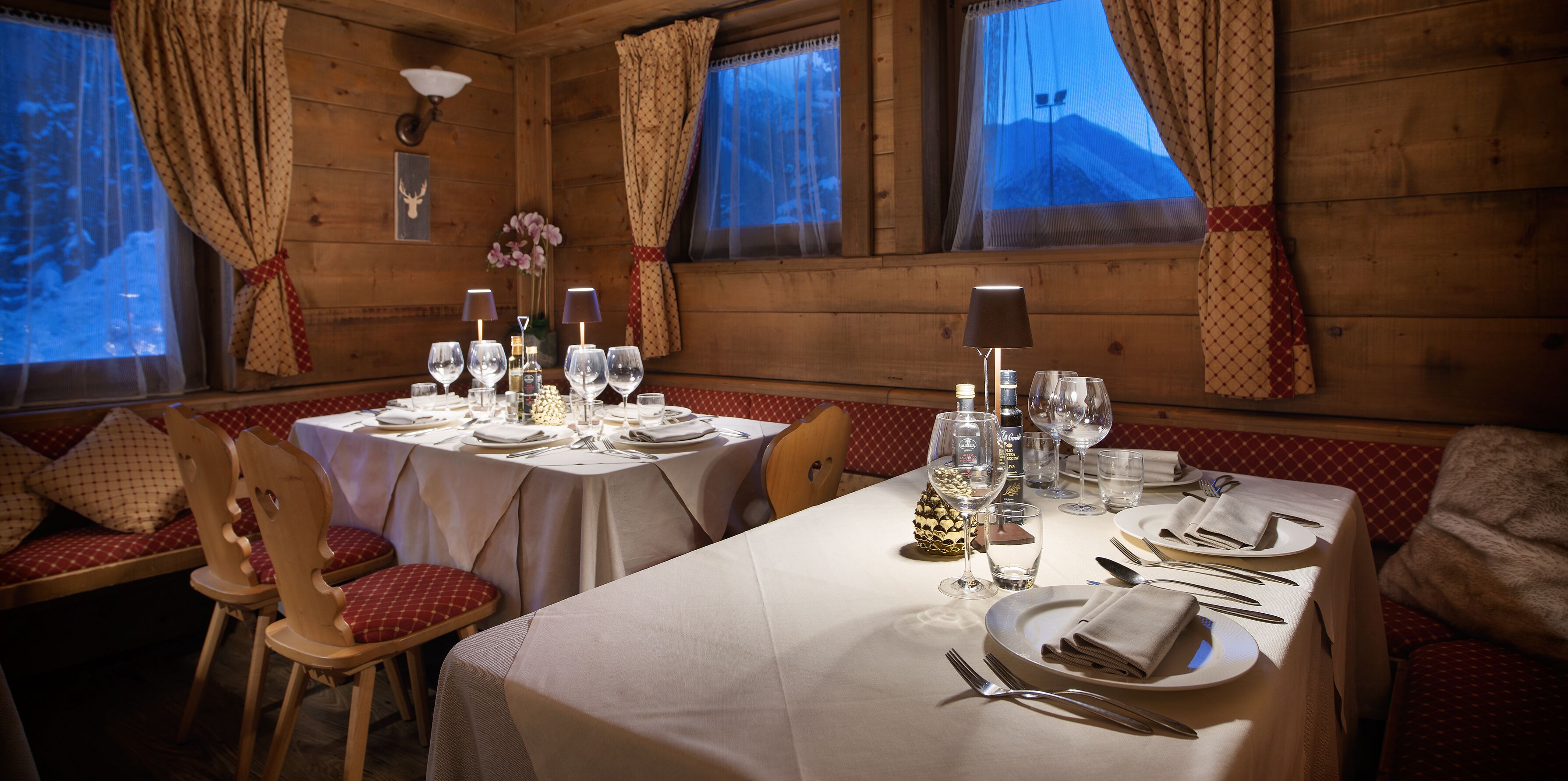 A cozy wooden cabin dining area with tables set for four, each with white tablecloths, red cushioned benches, and a variety of glassware and cutlery.