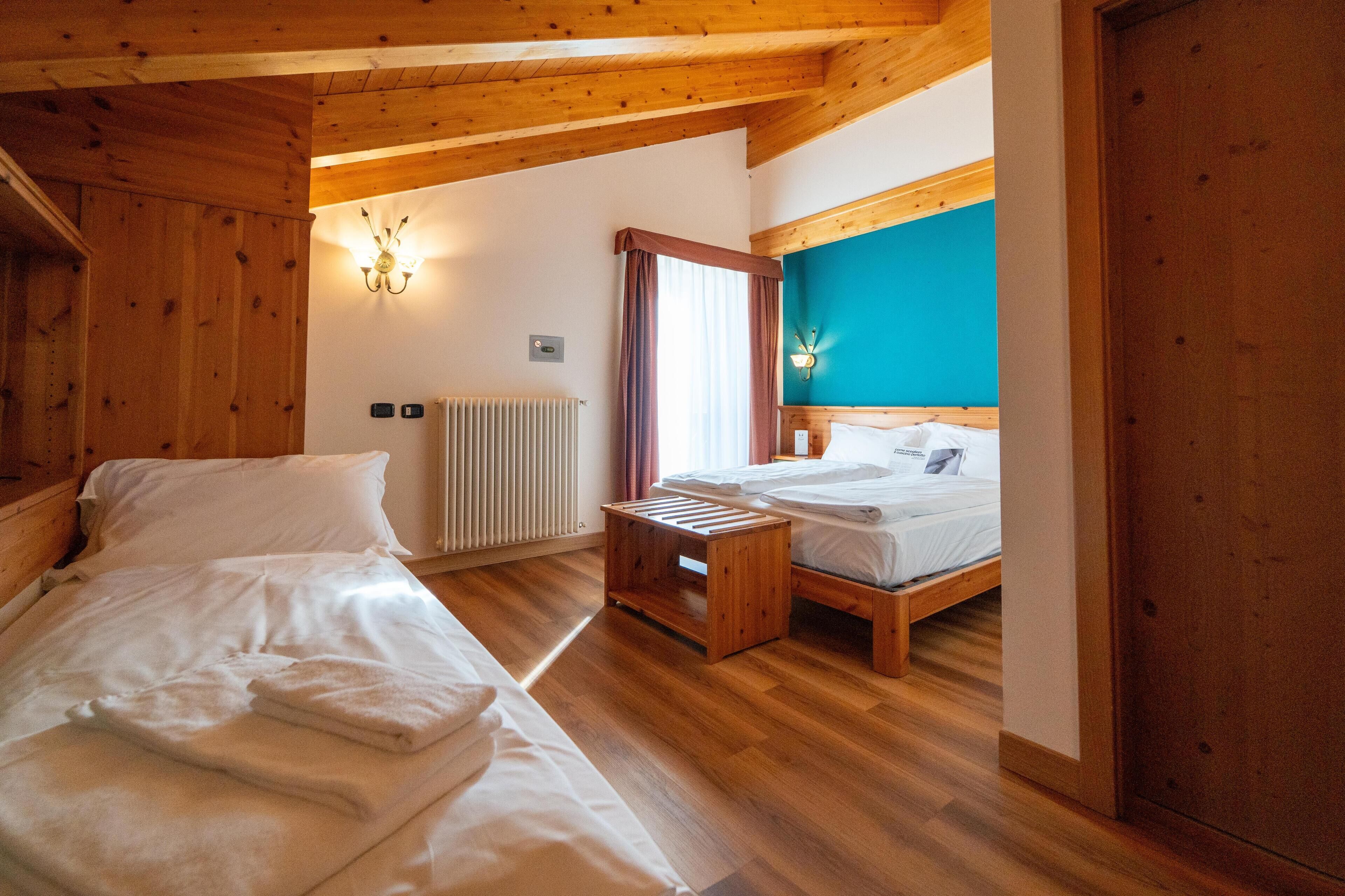 Una habitación con pisos de madera, una cama con sábanas blancas, un banco de madera, un aplique de pared y una ventana con cortinas.