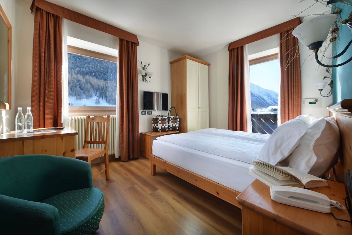Une chambre d’hôtel avec un lit en bois, un bureau avec une chaise, une télévision et une vue sur un paysage enneigé.