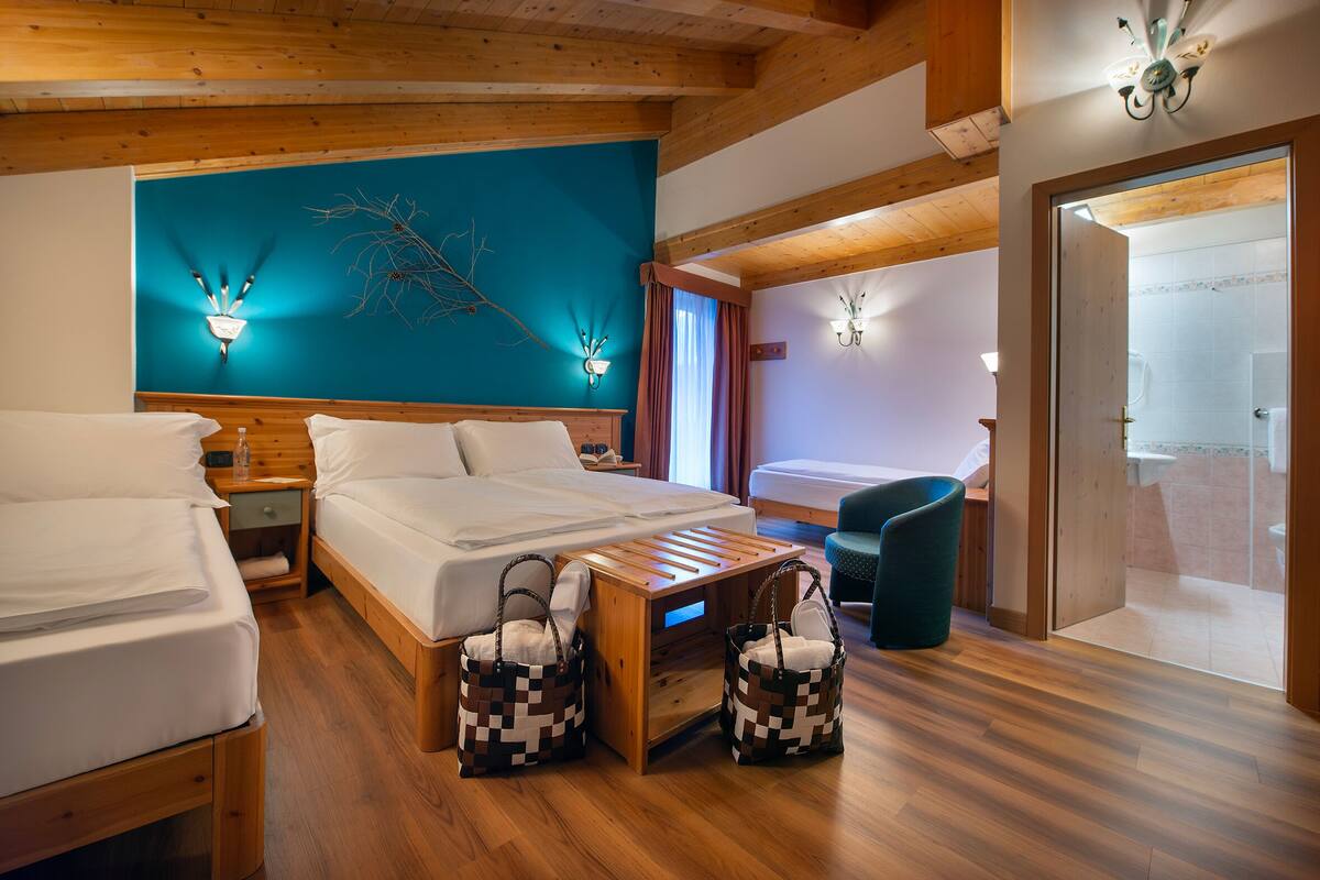 Une chambre d’hôtel avec un plancher en bois, un grand lit, deux tables de chevet, une petite table en bois, un luminaire fixé au mur, une petite table en bois, deux paniers en osier et une vue sur la salle de bain par une porte ouverte.