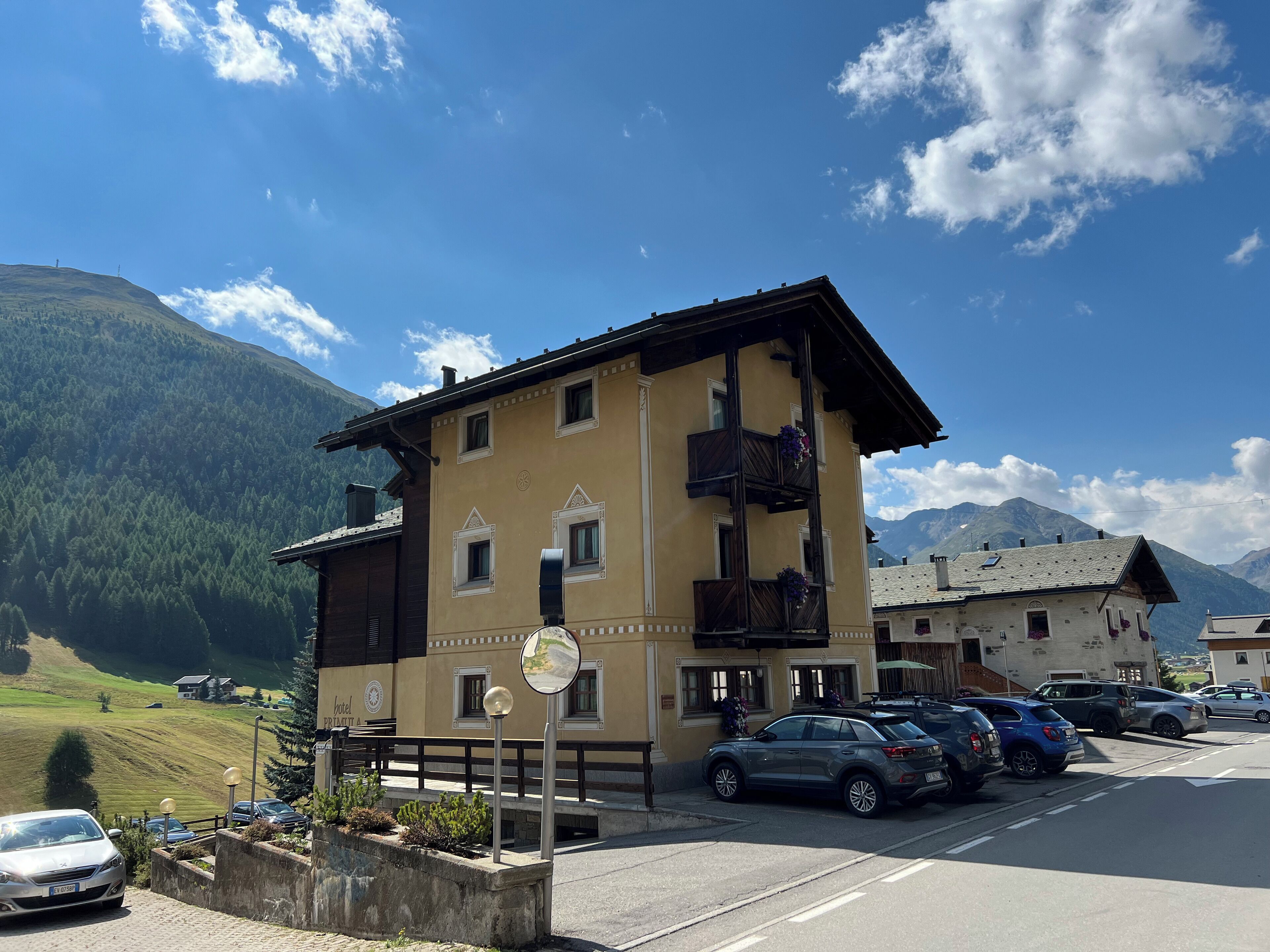 Ein zweistöckiges Gebäude mit Balkon, ein Parkplatz mit Autos und eine Bergkulisse im Hintergrund.
