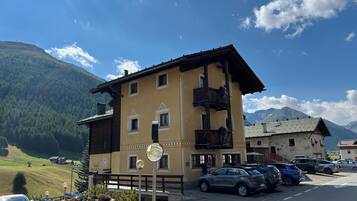 Ein zweistöckiges Gebäude mit Balkon, ein Parkplatz mit Autos und eine Bergkulisse im Hintergrund.
