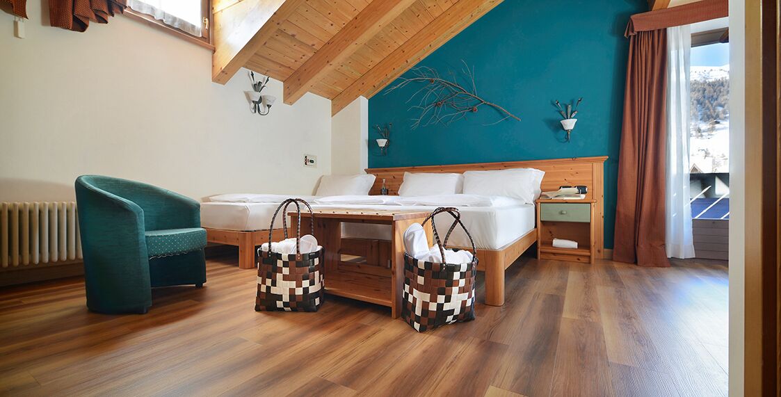 Una habitación con dos camas, una mesa de madera, un sillón azul turquesa y vistas a un paisaje nevado.