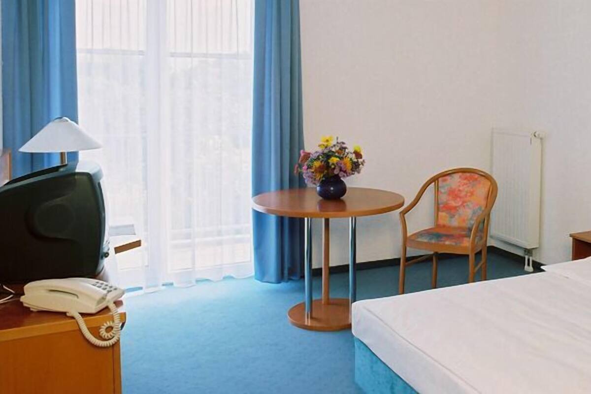 Une chambre d’hôtel avec un lit, un bureau avec un téléphone, une chaise, une petite table ronde ornée d’un vase de fleurs et un tableau accroché au mur.