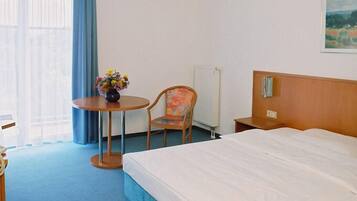 A hotel room with a bed, a desk with a telephone, a chair, a small table with a vase of flowers, and a painting on the wall.